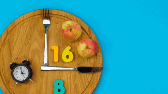 A wooden plate designed as a clock, with a fork and knife as clock hands pointing to numbers 16 and 8. Two apples are on the plate. A small black alarm clock is placed on the left. The background is bright blue.