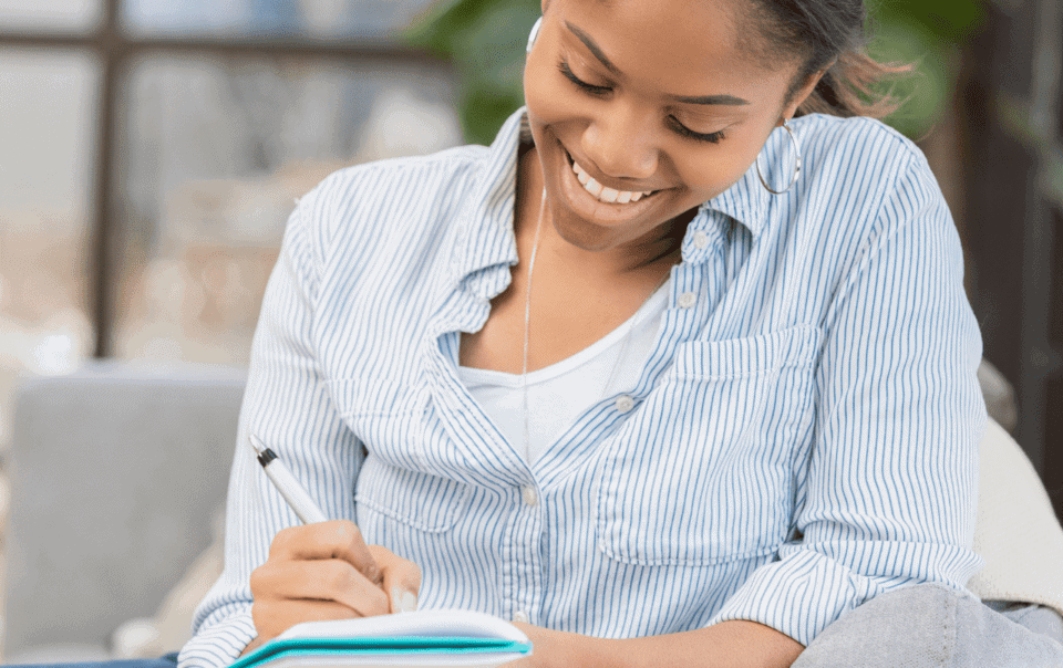 A smiling person in a striped shirt and earphones sits on a couch, jotting down weight loss goals in a notebook.