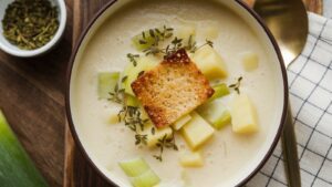 A bowl of creamy Potato Leek Soup garnished with diced potatoes, leeks, a toasted crouton, and fresh thyme sits beside a gold spoon and a checkered napkin on a wooden surface.