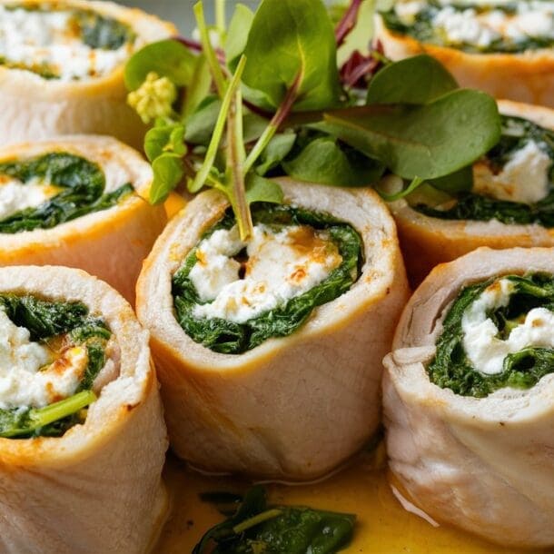 Cottage Cheese and Spinach Stuffed Chicken Roll-Ups are artfully arranged in a circle on a plate. Each rolled chicken breast is garnished with fresh greens and drizzled with a light sauce, creating an elegant and delicious presentation.