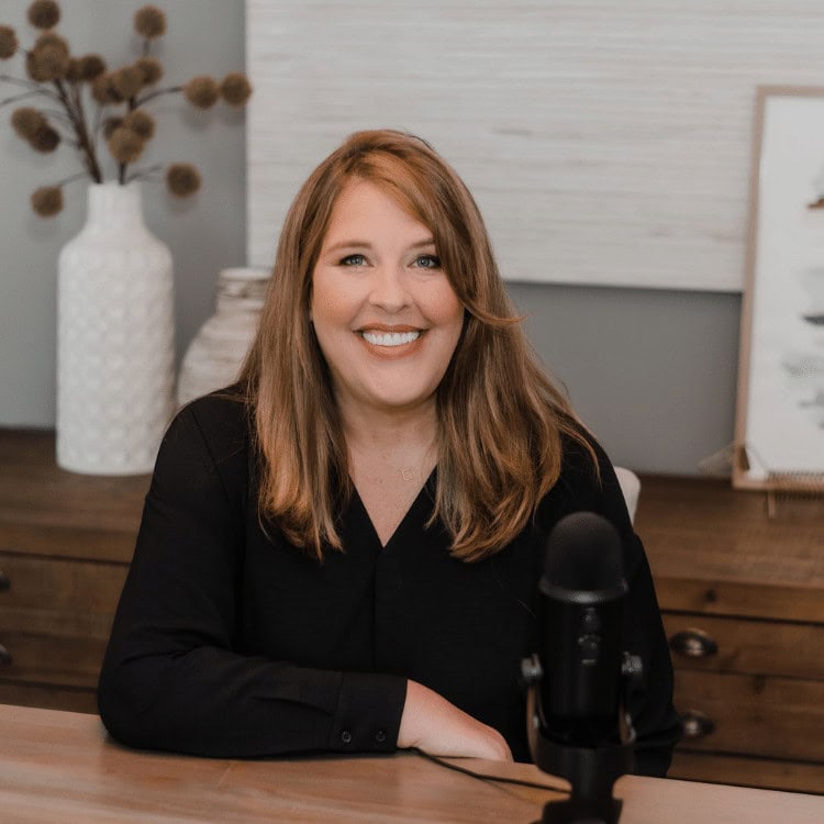 A woman with long brown hair and a black shirt sits at a wooden desk with a microphone, ready to share insights about eXp Realty. Behind her are a textured white vase with dried flowers and framed artwork. She smiles warmly at the camera, inviting engagement.