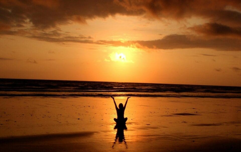 A person practices meditation for stress on a beach at sunset, arms raised and silhouetted against the orange sky reflected in wet sand. Waves roll in beneath dramatic clouds, creating a serene scene to find calm.