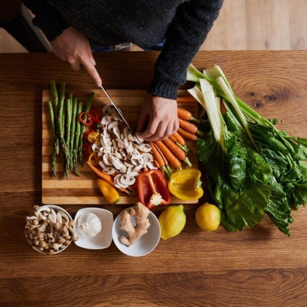 A person slices mushrooms on a cutting board surrounded by fresh vegetables, highlighting the nutrition found in asparagus, carrots, peppers, lettuce, lemons, ginger, and mushrooms on a wooden table.