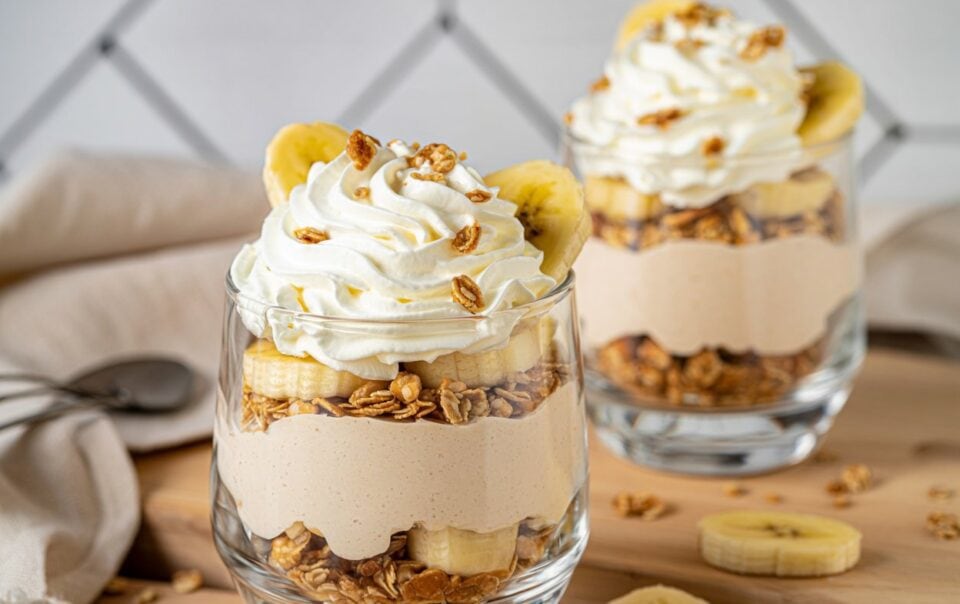 Two glass cups layered with granola, banana slices, creamy yogurt, and topped with whipped cream and granola. Sliced bananas and a gray napkin are visible on the wooden surface nearby.