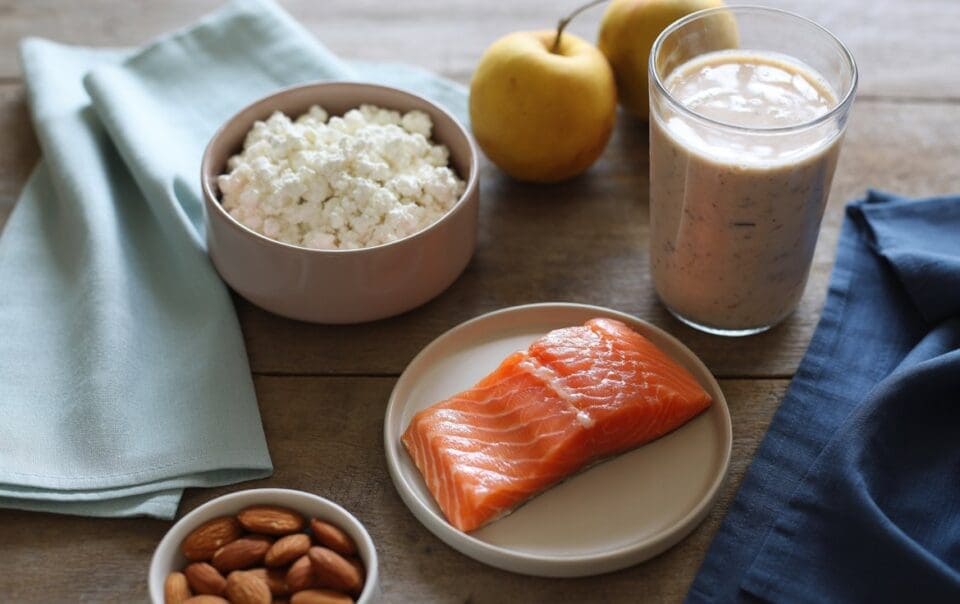 A wooden table displays a bowl of cottage cheese, a glass of smoothie, two yellow apples, a plate of raw salmon, and almonds—an ideal spread if you’re wondering how much protein should you eat daily. Light blue and navy napkins sit nearby.