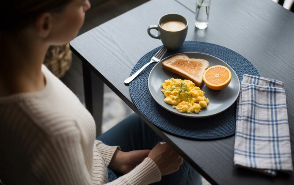 A person sits at a table with a plate of scrambled eggs, toast, and half an orange—natural ways to feel full—next to a mug of coffee and a glass of water. A fork and a blue-checked napkin are on the table.