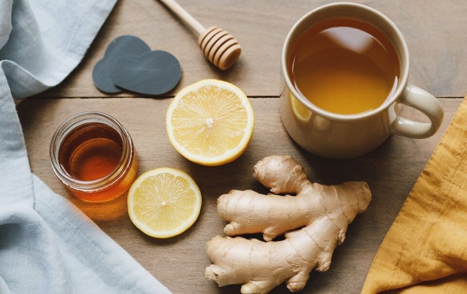 A mug of tea, a jar of honey with a honey dipper, fresh ginger root, and sliced lemon are arranged on a wooden table with blue and yellow cloth napkins—classic staples for natural cold remedies.