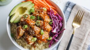 A Protein Bowl featuring quinoa, Spicy Honey Chicken glazed and topped with sesame seeds and green onions, sliced avocado, shredded carrots, and purple cabbage, served with a fork and napkin on the side.