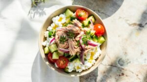 A Tuna Cottage Cheese Power Bowl filled with cucumber chunks, cherry tomato halves, red onion slices, and flaked tuna, garnished with fresh herbs, sits on a light surface with sunlight casting soft shadows.