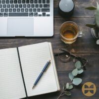 A wooden desk with an open notebook, pen, eyeglasses, a cup of tea, a laptop, a small speaker, and plant leaves. The gold circular logo in the bottom right corner reflects the WellNest Guide Editorial Team’s attention to detail.