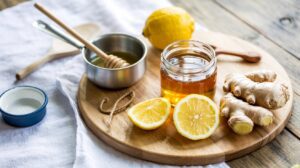A wooden board with a jar of honey, sliced lemon, whole lemon, fresh ginger root, and a small bowl of honey with a dipper—classic ingredients for DIY Natural Cough Syrup—arranged on a white cloth on a rustic wooden table.