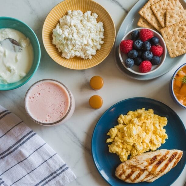 A breakfast spread featuring scrambled eggs, grilled chicken, cottage cheese, yogurt, a bowl of berries, crackers, a cup of pink smoothie, soup, and a striped napkin on a marble surface offers delicious food swaps for Wegovy users.