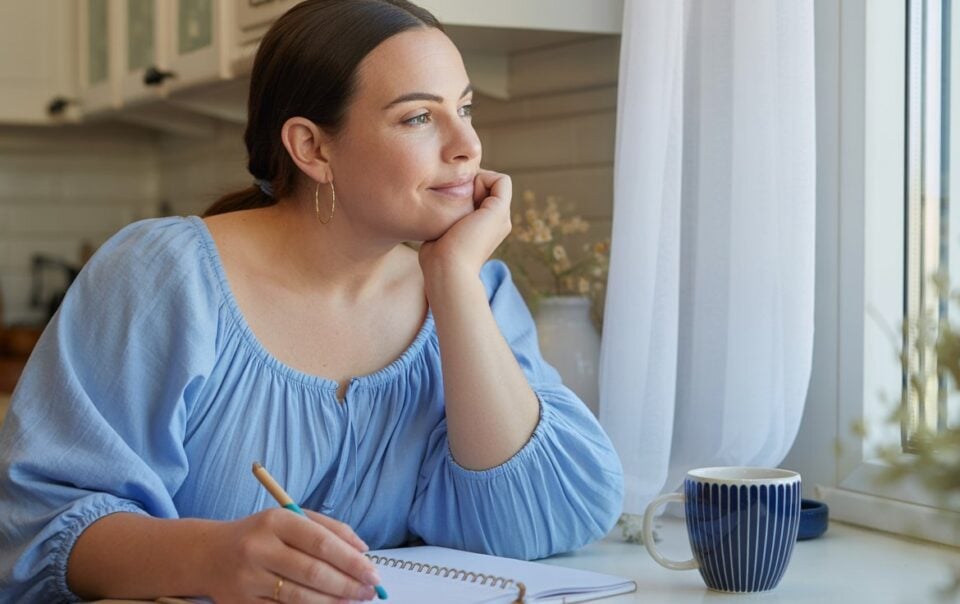 A woman in a light blue blouse sits at a kitchen table, resting her chin on her hand and looking thoughtfully out the window. Reflecting on her 110 pounds weight loss journey, she holds a pen over an open notebook, with a striped mug nearby.