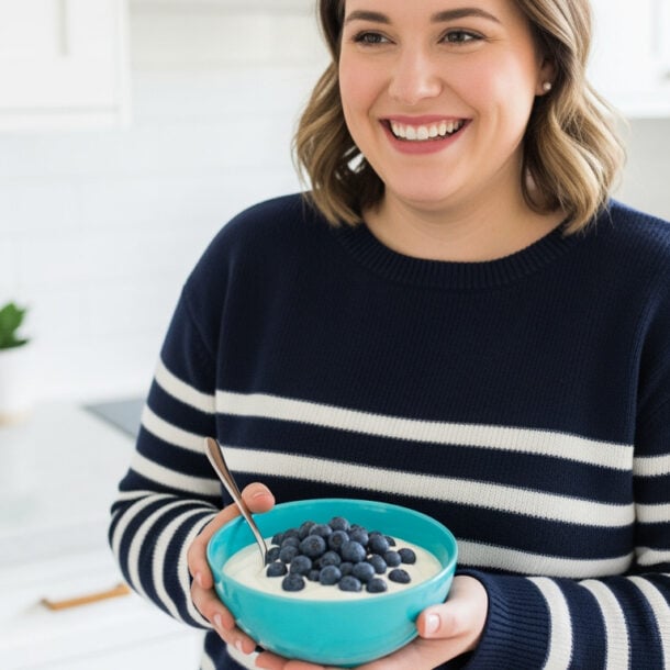 A smiling woman in a navy and white striped sweater holds a bowl of yogurt topped with fresh blueberries, enjoying a bright, modern kitchen as she follows the 30-30-30 Rule for a balanced morning routine.