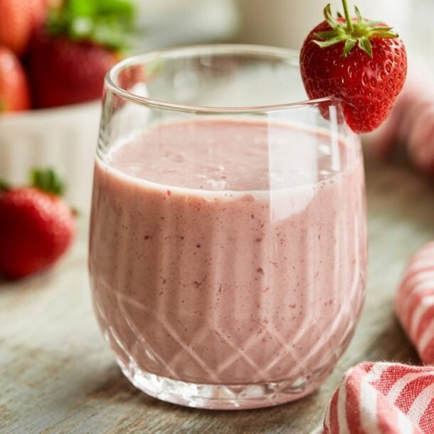 A glass of pink cottage cheese smoothie, high in protein, is garnished with a whole strawberry on the rim. It sits on a wooden table next to a red and white striped cloth and a bowl of fresh strawberries.