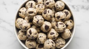 A bowl filled with round cookies and cream protein balls mixed with chunks of chocolate, placed on a white marble surface.