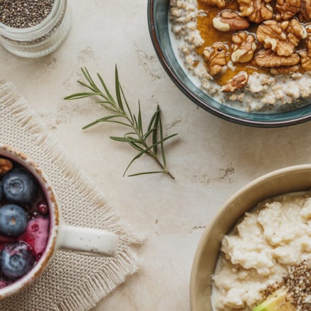 Three bowls on a table showcase breakfast recipes perfect for a cortisol reset: oatmeal with walnuts and honey, oatmeal with blueberries and walnuts, and oatmeal with avocado slices and seeds. A small jar of chia seeds and a rosemary sprig are nearby.