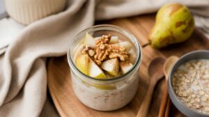 A jar of High Protein Pear Walnut Overnight Oats topped with sliced pears and walnuts sits on a wooden tray. Nearby are a whole pear, a bowl of oats, wooden spoons, and a beige cloth napkin.
