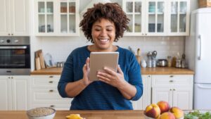 A woman stands in a bright kitchen, smiling while using a tablet. Fresh fruit and estrogen-rich foods are on the counter in front of her, supporting women's health. White cabinets and kitchenware are in the background.