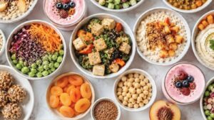 An assortment of colorful, healthy dishes in bowls, featuring hormone balancing recipes like salads with tofu, chickpeas, edamame, chia puddings with fruit, dried apricots, smoothie bowls, energy bites, and sliced peaches on a light surface.