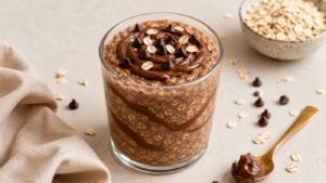 A glass filled with Nutella overnight oats, topped with chocolate chips and oats. A gold spoon with a chocolate dollop rests nearby, with a bowl of high protein oats and scattered chocolate chips on a light surface.
