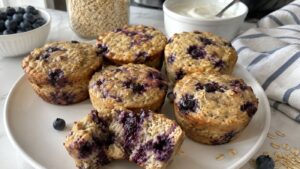 Six blueberry muffins on a white plate, one muffin broken open to show the inside. Reminiscent of blueberry cheesecake, these high-protein baked oatmeal cups are surrounded by fresh blueberries, oats, yogurt, and a striped napkin on the marble countertop.
