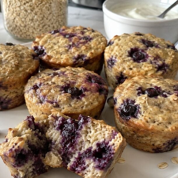 Six blueberry muffins on a white plate, one muffin broken open to show the inside. Reminiscent of blueberry cheesecake, these high-protein baked oatmeal cups are surrounded by fresh blueberries, oats, yogurt, and a striped napkin on the marble countertop.