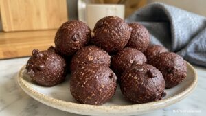 A plate filled with round chocolate energy bites dotted with chocolate chips sits on a marble countertop with a blurred background of a mug, cutting board, and gray cloth.