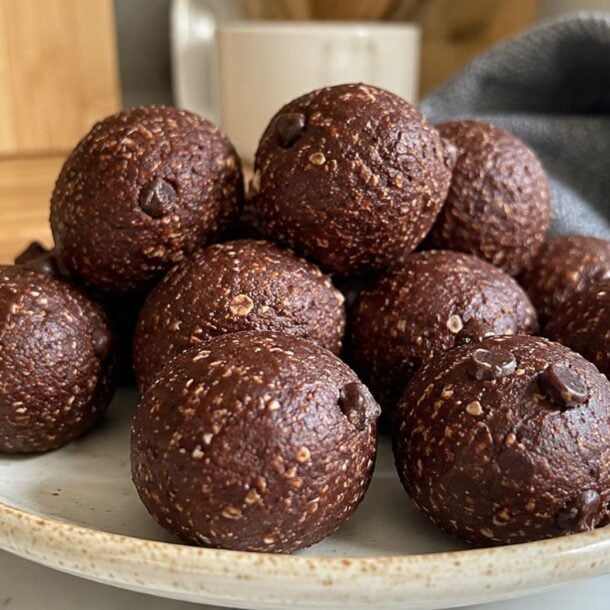 A plate filled with round chocolate energy bites dotted with chocolate chips sits on a marble countertop with a blurred background of a mug, cutting board, and gray cloth.