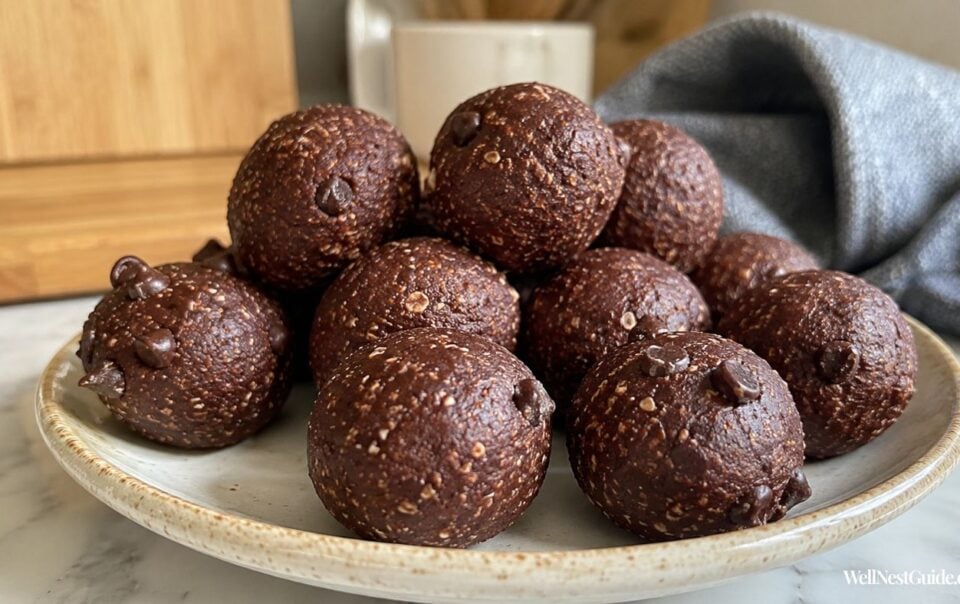 A plate filled with round chocolate energy bites dotted with chocolate chips sits on a marble countertop with a blurred background of a mug, cutting board, and gray cloth.