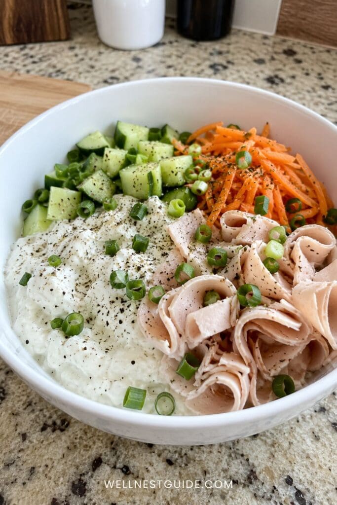 A white bowl filled with cucumber cubes, shredded carrots, thinly sliced turkey, and cottage cheese topped with black pepper and green onions—a refreshing no-cook meal set on a speckled kitchen countertop.