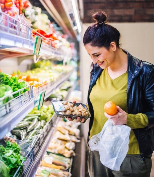 A woman smiles while grocery shopping, holding a tomato and a plastic-wrapped pack of mushrooms—perfect ingredients for GLP-1 recipes—in the produce section filled with fresh vegetables.